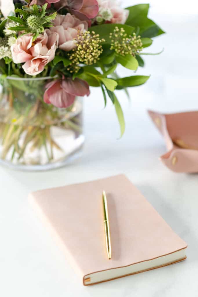A soft pink journal with a gold pen placed on top, next to a vase of flowers, used on the Print and Hustle homepage to navigate to the budget and save planners category.