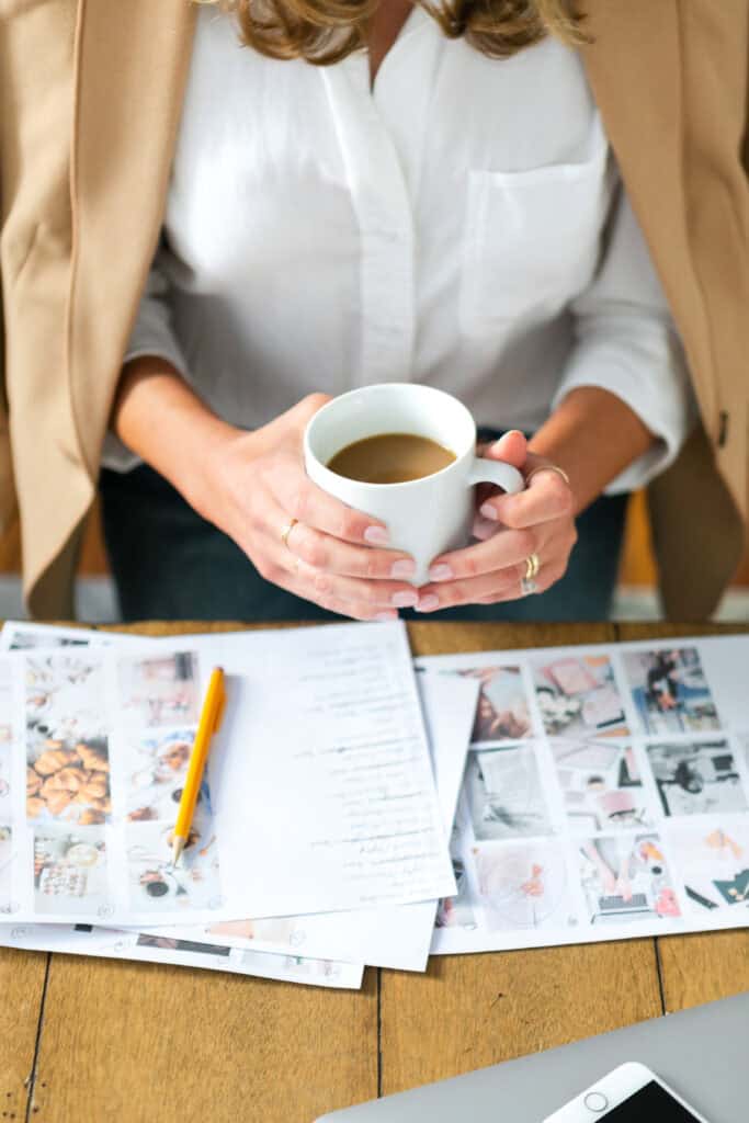 A woman holding a coffee cup while sitting at a desk with planning documents and images, used on the Print and Hustle homepage to navigate to the lifestyle planners category.