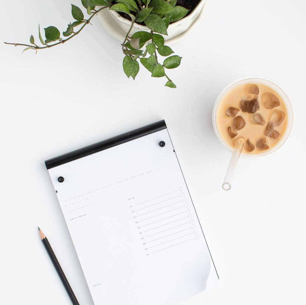 A notepad with a to-do list layout next to a pencil, a cup of iced coffee, and a potted plant, representing the organization and planning involved in a family chore chart for busy moms.