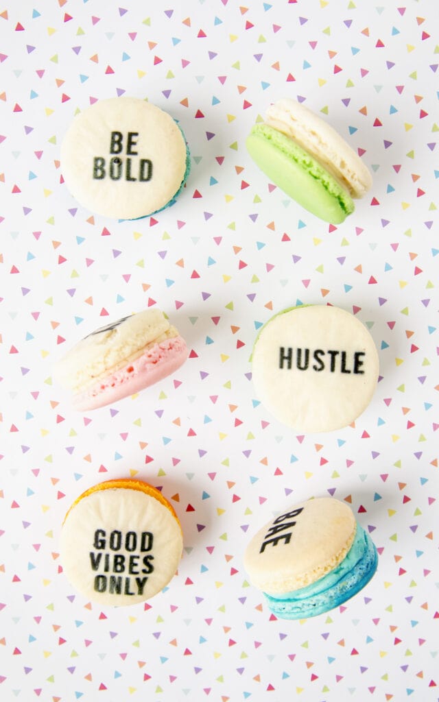 Colorful macarons with motivational words like "Be Bold," "Hustle," "Good Vibes Only," and "Bae" on a playful, confetti-patterned background, used on a website homepage to navigate to the printables section