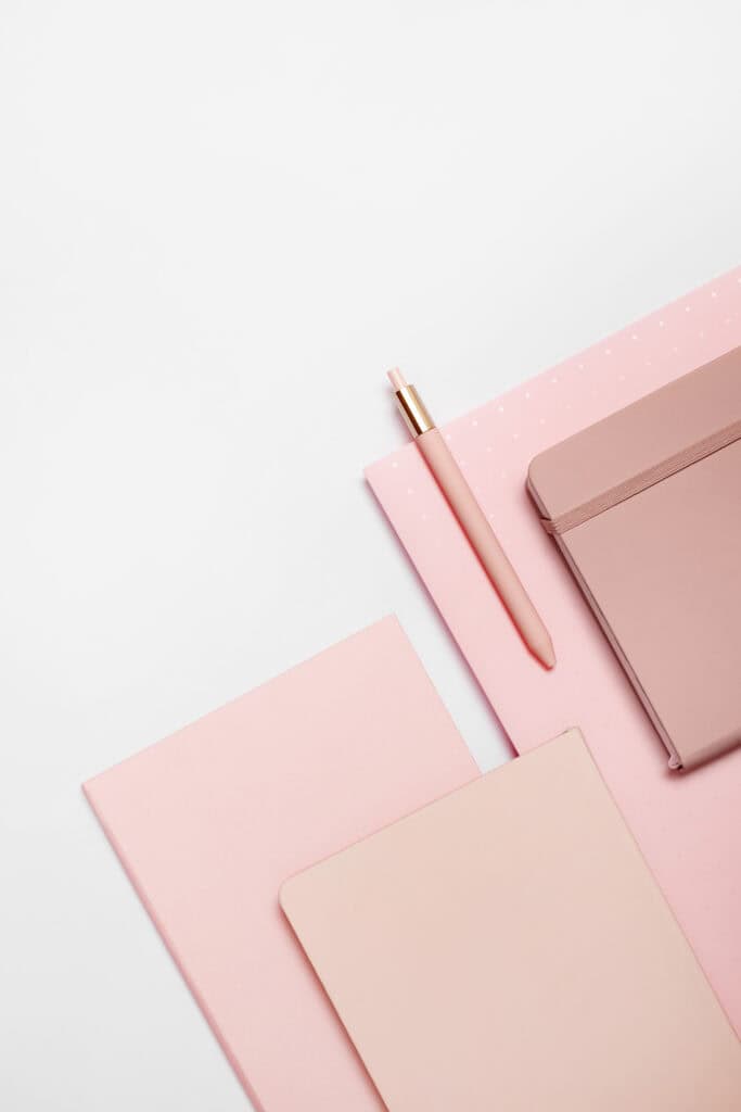Flat lay of pink and blush-colored notebooks and a pen, used on the Print and Hustle homepage to navigate to the business printables category.