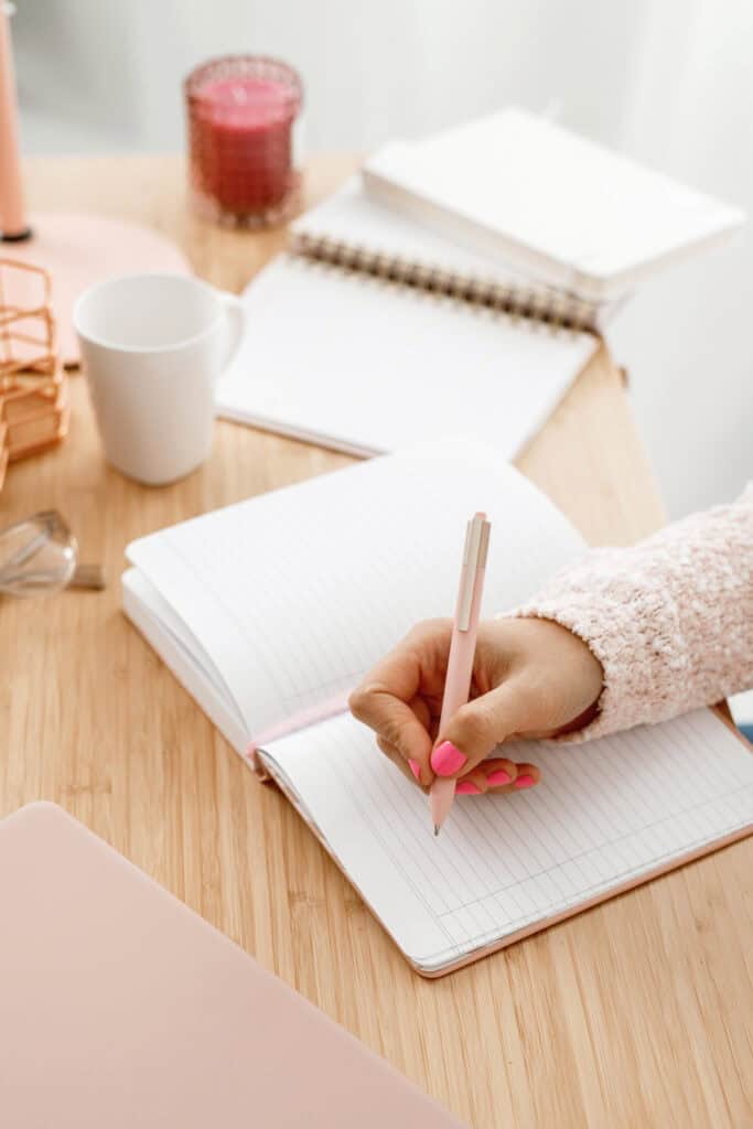 Feminine workspace with a blush pink aesthetic featuring a cozy desk setup, an open notebook, and a hand writing with a pink pen, surrounded by a coffee mug, candle, and accessories for productivity and creativity.