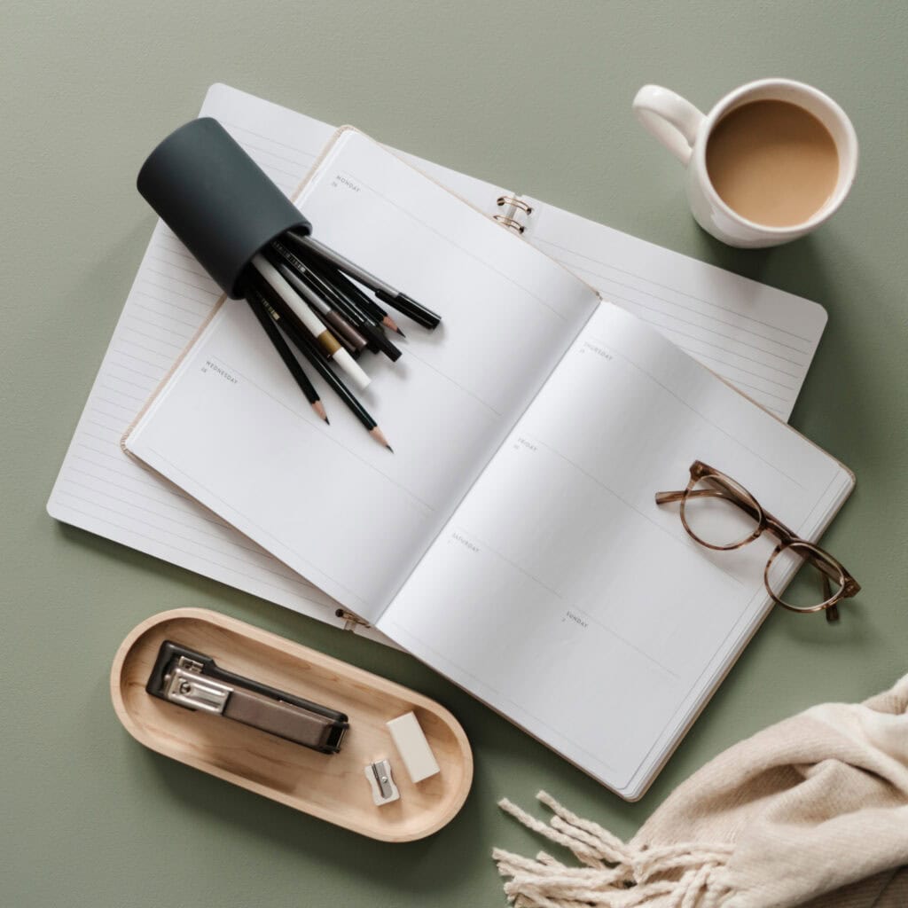 An open planner with blank weekly pages, surrounded by pencils, a pair of glasses, a coffee mug, and office supplies, symbolizing the process of choosing the right planner for managing tasks effectively.