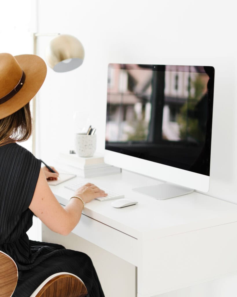 A mom wearing a brown hat works at a clean, white desk in a bright home office, typing on a computer, symbolizing organized family life and planning.
