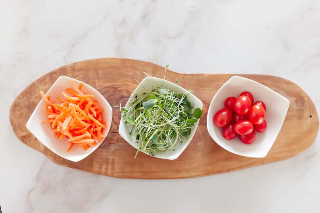 Three small white bowls with shredded carrots, fresh green sprouts, and cherry tomatoes, placed on a wooden board, illustrating healthy meal prep for a weekly meal planner printable designed for moms.
