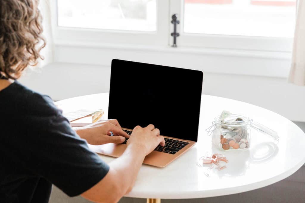 A person typing on a laptop at a round table with a jar of coins and dollar bills nearby, illustrating the use of family budgeting printables to simplify financial planning for busy moms.