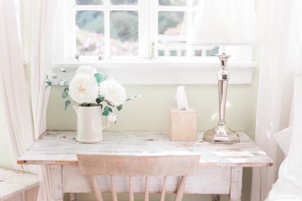  A serene desk by a window with a vase of white flowers, a box of tissues, and a silver lamp, symbolizing a calm and reflective space ideal for moms to use gratitude planners