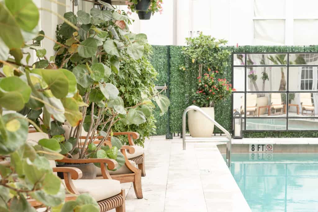 A serene poolside area with lush green plants and wooden chairs, offering a tranquil setting. This image is featured in a post on printandhustle.com about the best flexible side hustles for stay-at-home moms.