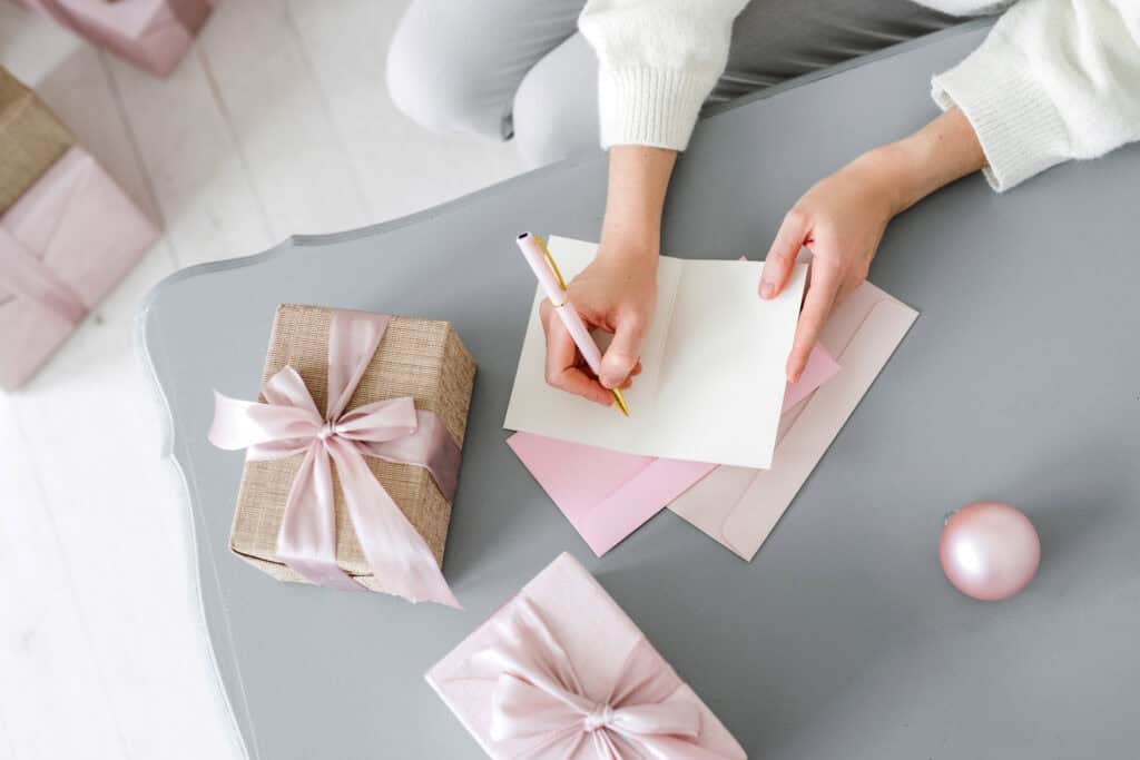 A person sits at a table writing a card, surrounded by pink and beige gift boxes tied with pink ribbons. Pink envelopes and a Christmas ornament are also on the table. This image represents holiday and event planning, emphasizing the use of printables to simplify tasks like gift preparation and card writing for busy moms.