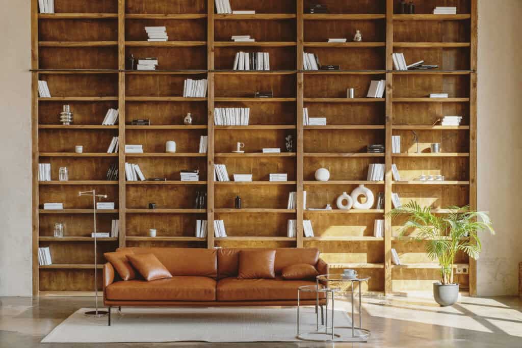 A modern living room with a large wooden bookshelf filled with books and decorative items, a brown leather sofa, and a small side table, illustrating a well-organized household space ideal for effective management by moms.