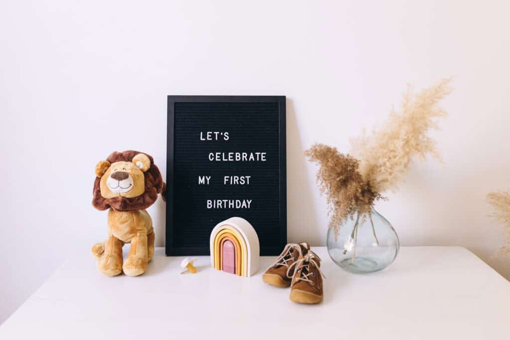 A display with a felt board that reads 'Let's celebrate my first birthday,' accompanied by a stuffed lion, a wooden rainbow stacker, baby shoes, and a vase with pampas grass, illustrating birthday party planning printables for busy moms.