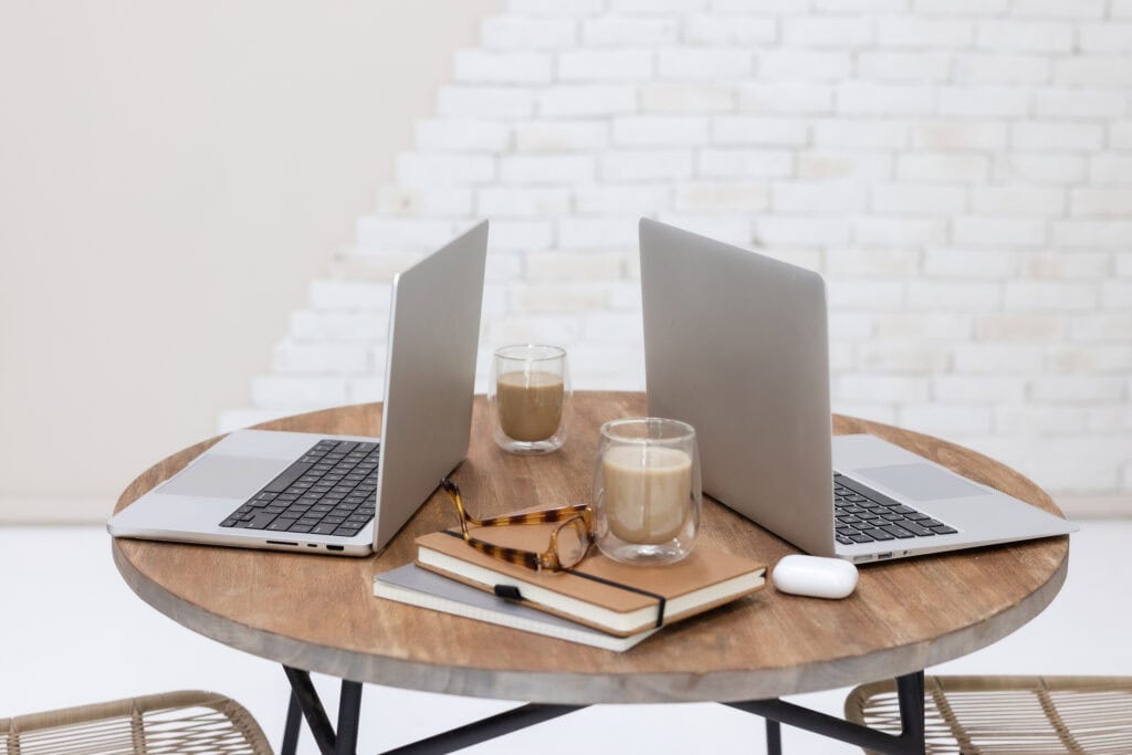 Two laptops on a round wooden table with notebooks, glasses, and coffee cups, illustrating a workspace setup. This image is featured in a post on printandhustle.com about the best flexible side hustles for stay-at-home moms, highlighting the potential of freelancing and virtual assistant work