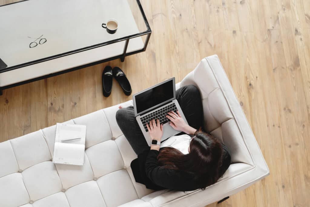 Overhead view of a woman working on a laptop while sitting on a white sofa, with a notebook and a coffee cup nearby. This image is featured in a post on printandhustle.com about the best flexible side hustles for stay-at-home moms.