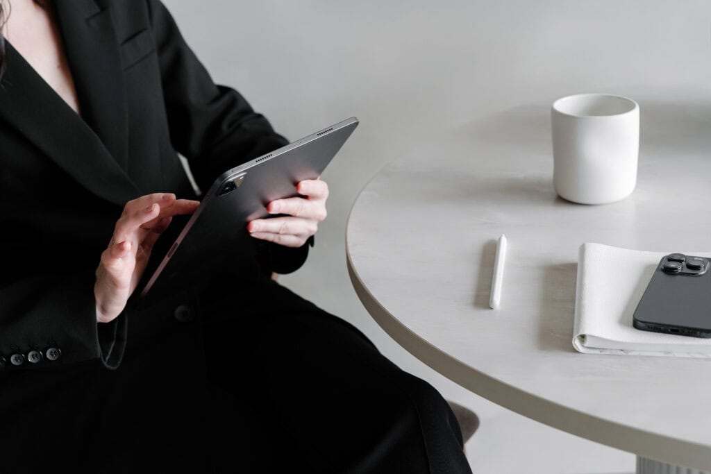 A person dressed in a black blazer uses a tablet at a minimalist table. On the table are a white mug, a stylus, a smartphone, and a white notebook. This image represents home business planning, highlighting the use of printables to help busy moms organize and manage their business ideas efficiently.