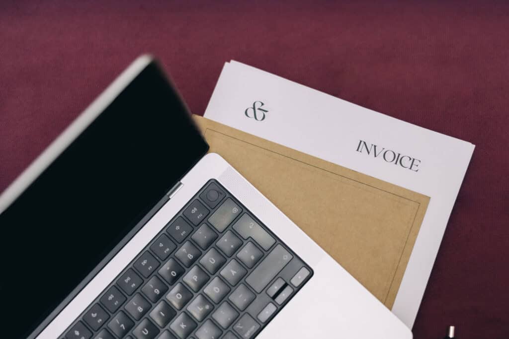 A laptop partially covering an invoice and other documents on a burgundy surface, representing the use of family budgeting printables to manage finances and simplify life for busy moms.