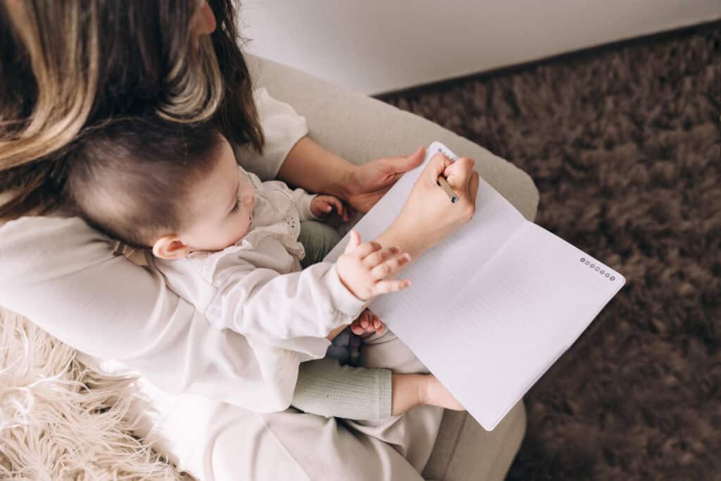 A mother holding her baby while writing in a notebook, illustrating the benefits of using printables to help busy moms stay organized