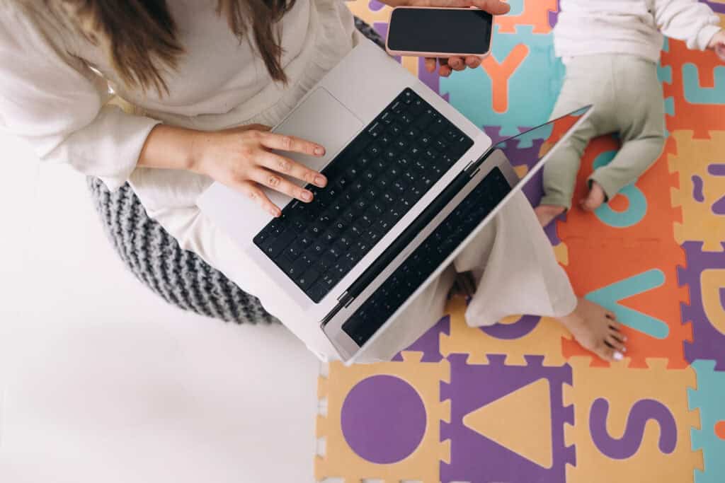 A stay-at-home mom working on a laptop while holding a smartphone, with a baby playing on a colorful mat nearby, illustrating the concept of flexible side hustles for stay-at-home moms, featured on printandhustle.com.