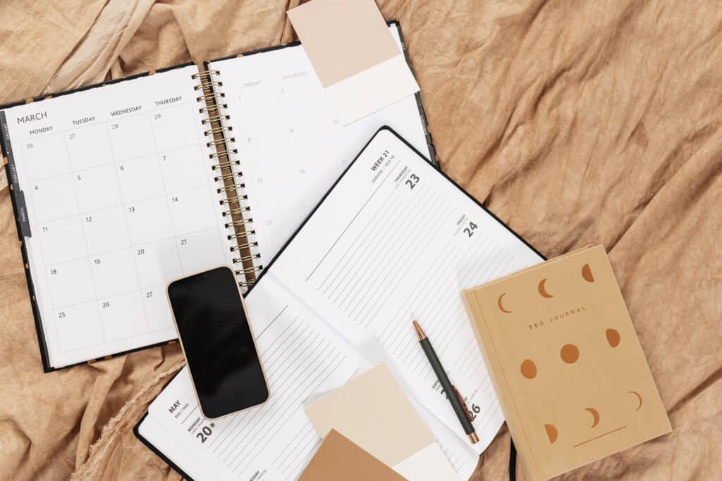 A flat lay of a calendar, an open weekly planner, a 365 journal, a smartphone, and color swatches arranged on a beige fabric. A pen rests on the planner, highlighting a focus on organization and scheduling. This image represents appointment tracker printables, illustrating tools busy moms can use to keep track of their schedules and appointments effectively.