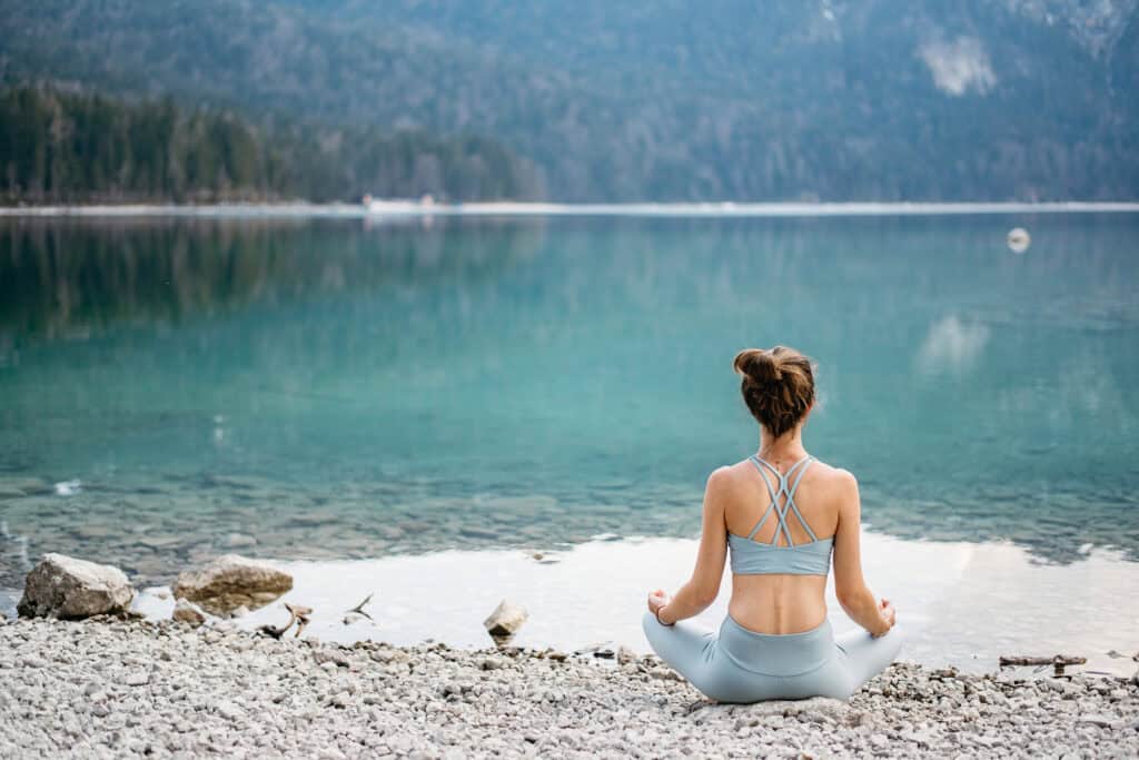 A woman sitting in a meditative pose by a serene lake, surrounded by mountains, representing the importance of self-care printables for busy moms to maintain a balanced and simpler life.
