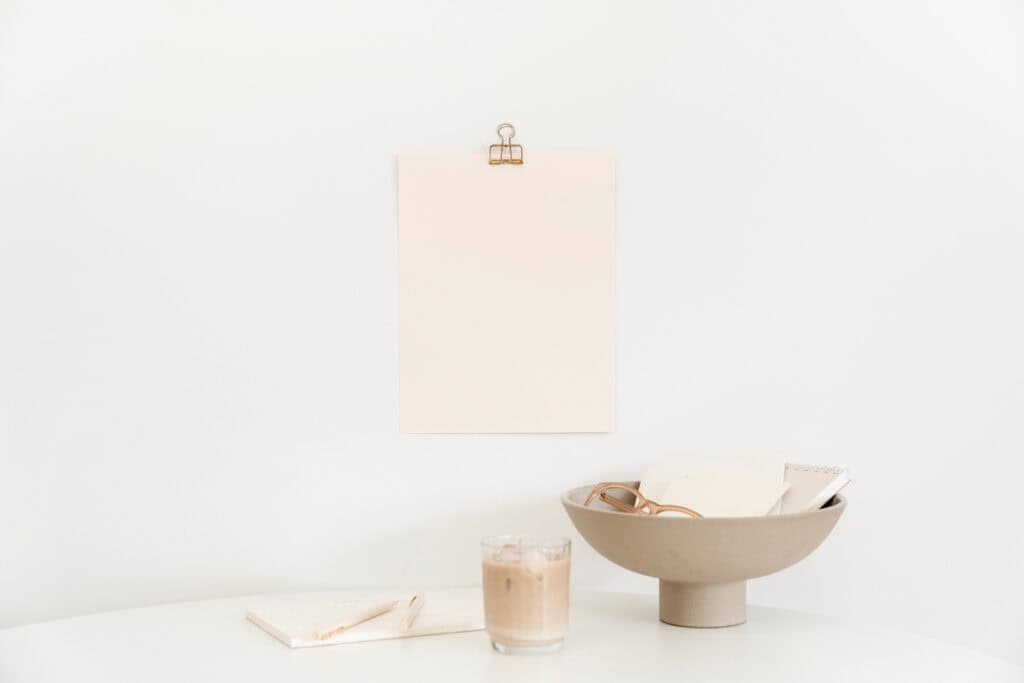 A minimalist setup featuring a blank sheet of paper clipped to a wall, a bowl with glasses and notepads, and a glass of iced coffee on a white table, representing the simplicity and organization of using daily schedules for kids.