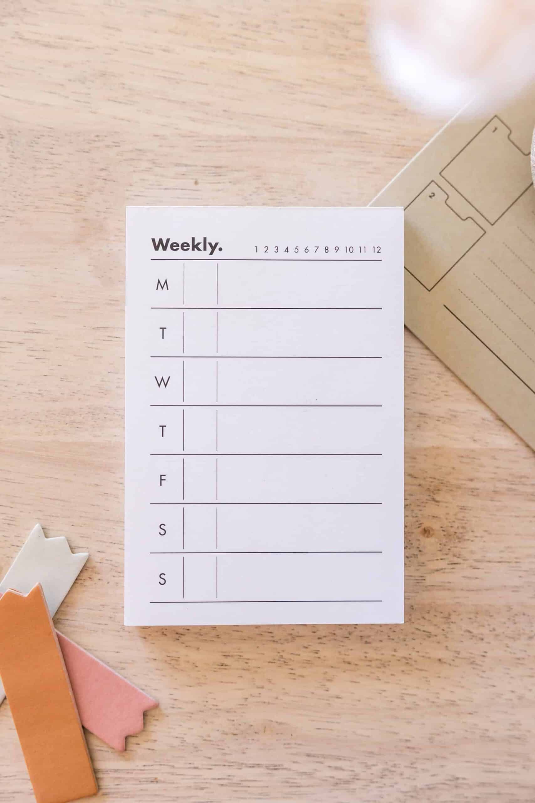 Minimalist weekly planner notepad placed on a wooden desk, accompanied by colorful page markers and an envelope, representing organization and productivity. Perfect for showcasing organizational printables for moms to stay productive in managing home, business, and family tasks. Image credit: Styled Stock Society.