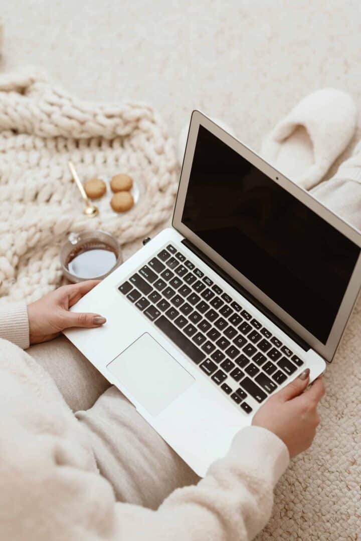 A cozy workspace setup with a person working on a laptop, surrounded by a chunky knit blanket, a cup of coffee, and cookies on a gold spoon—perfect for productive yet relaxed side hustle planning.
