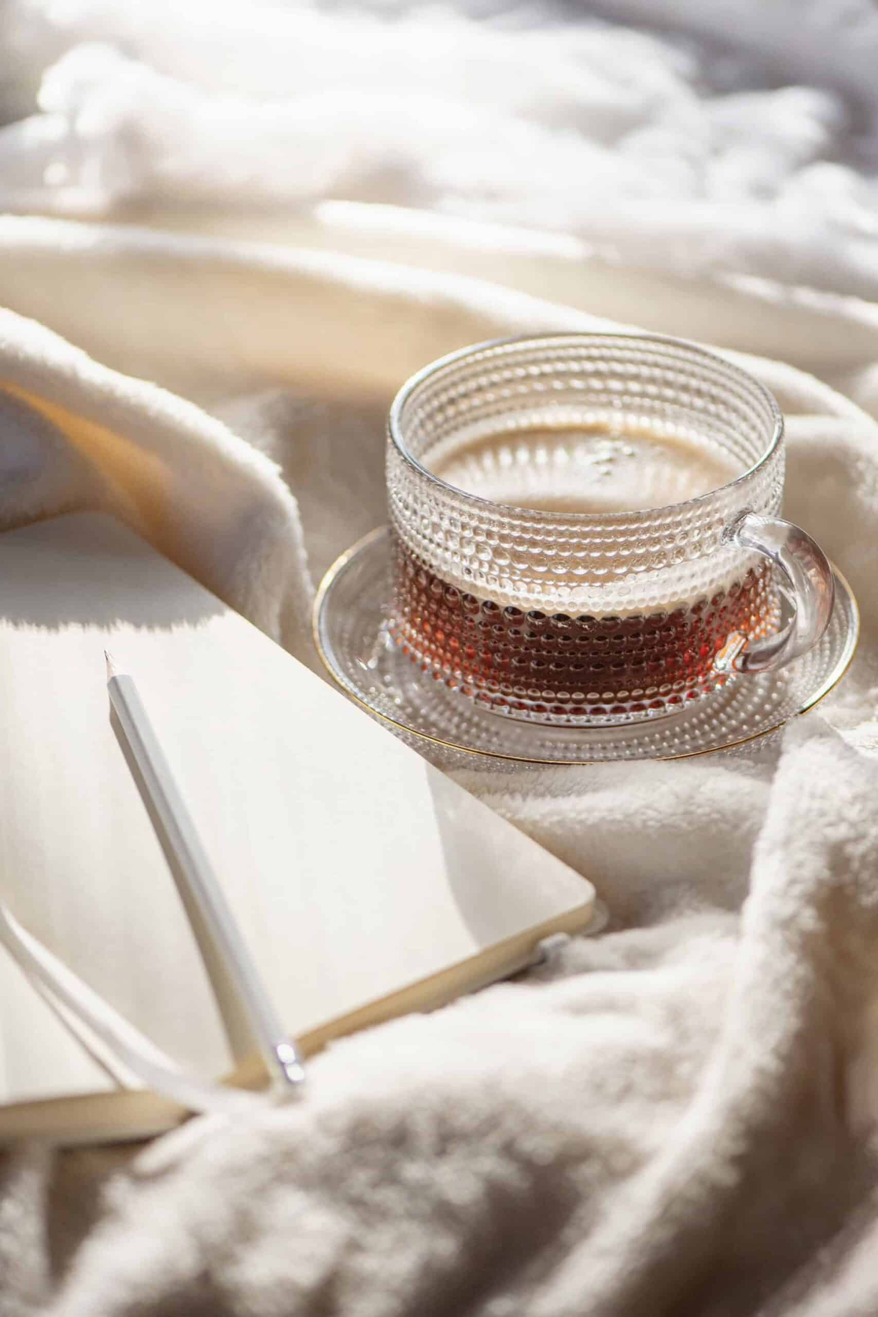 Cozy workspace setup with a glass cup of tea, an open notebook, and a pencil on a soft cream blanket, symbolizing calm and focused planning for starting a side hustle.