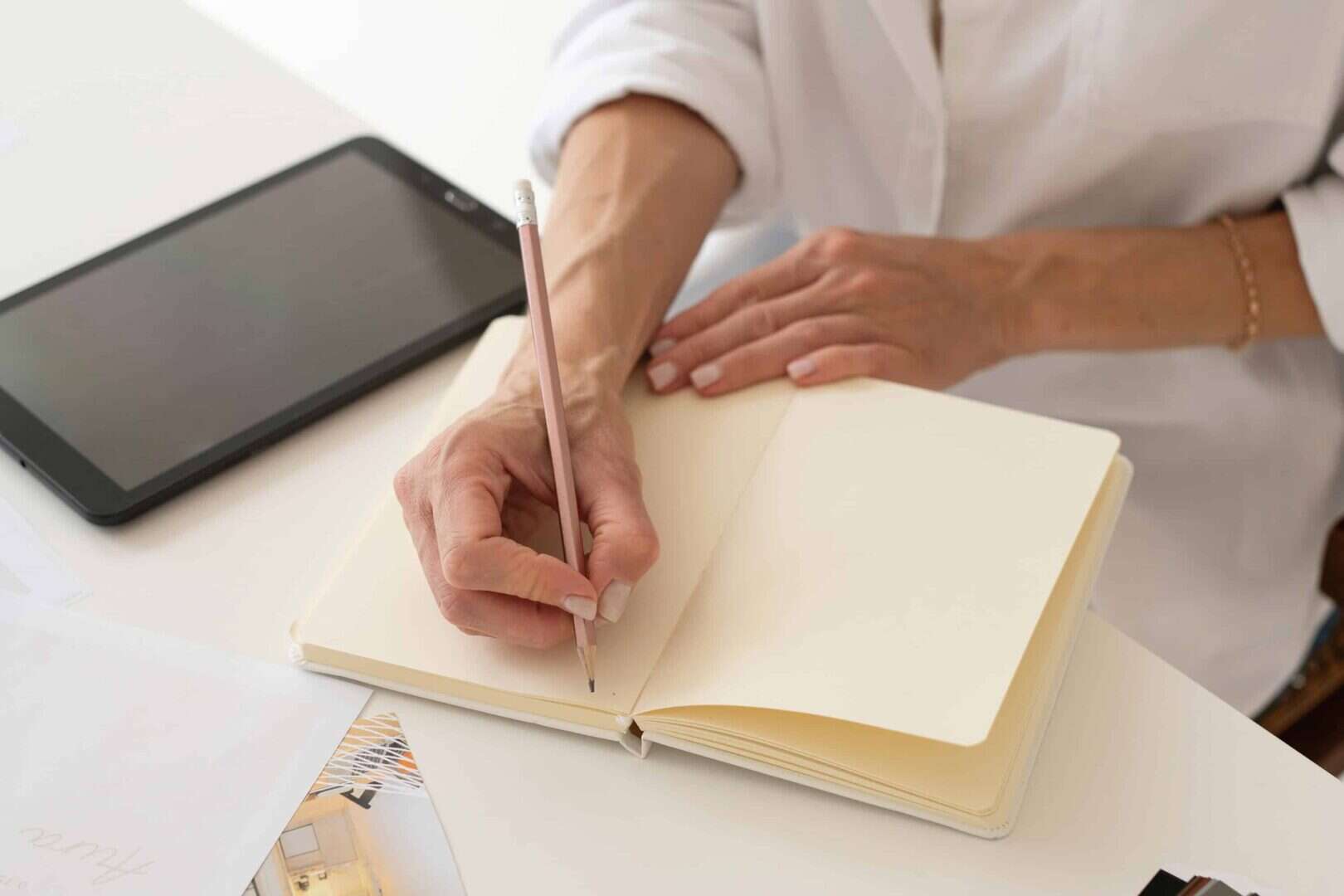 Close-up of a person writing in a paper planner with a pencil beside a tablet on a desk. Represents the benefits of a hybrid planner system that combines digital and printable planners for better organization.