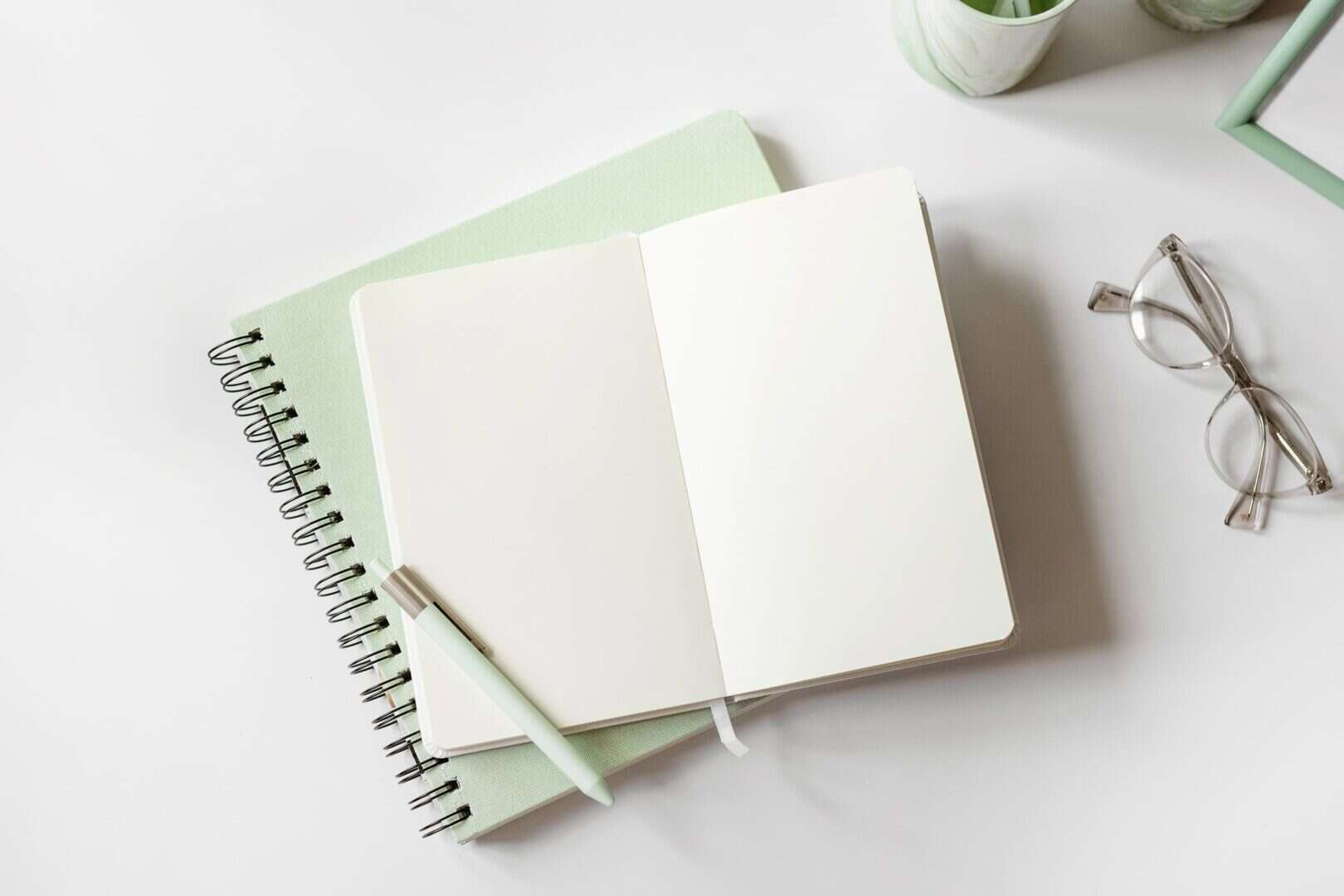Open blank notebook with a mint green pen on top of spiral planners, with eyeglasses nearby on a white desk. Represents starting a daily planner routine and building habits that stick.