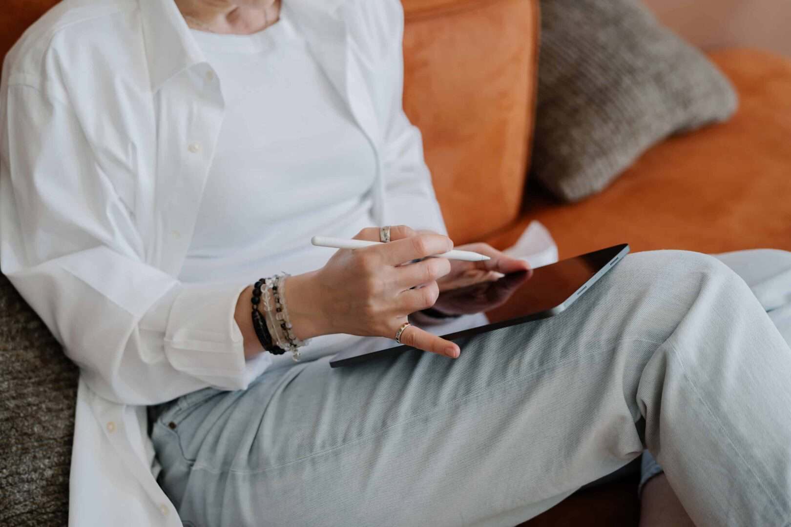 Woman sitting on a couch using a digital planner on a tablet with a stylus, wearing casual clothing and layered bracelets. Captures how creative entrepreneurs can stay organized with digital planner systems that support flexible, on-the-go productivity.