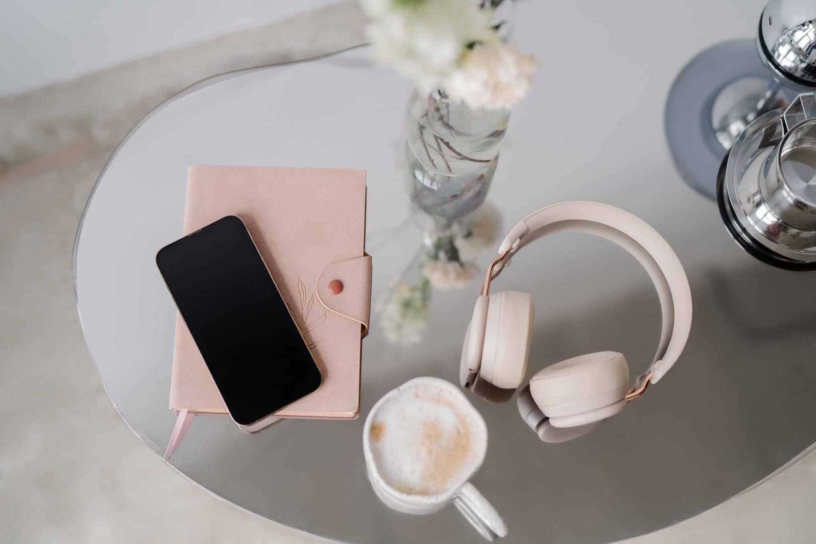 Flat lay of a smartphone resting on a blush pink planner, alongside cream-colored headphones, a latte, and a vase of flowers on a glass table. Highlights the relaxed yet efficient setup often used for digital planner systems by entrepreneurs managing tasks on the go.