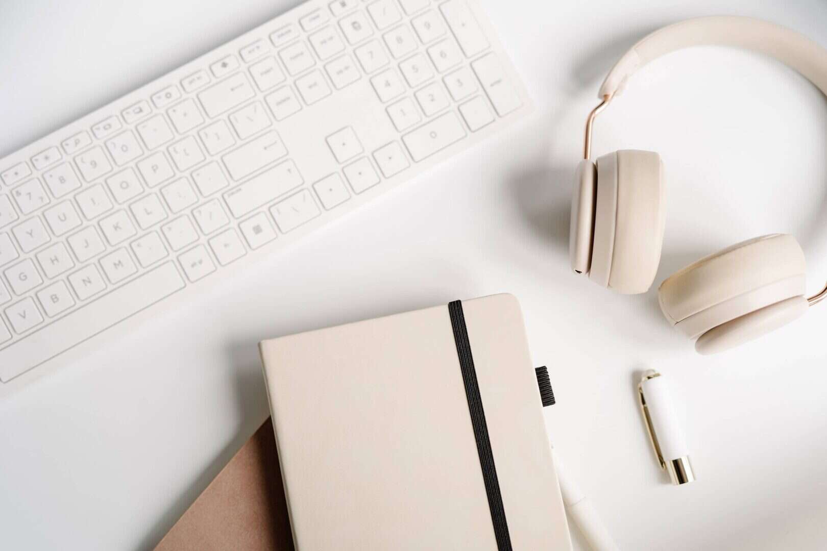 Flat lay of a beige notebook with a black elastic band, over-ear headphones, a white pen, and a wireless keyboard on a white desk. Represents a hybrid planner system that combines digital tools and printable planners for flexible productivity and workspace organization.