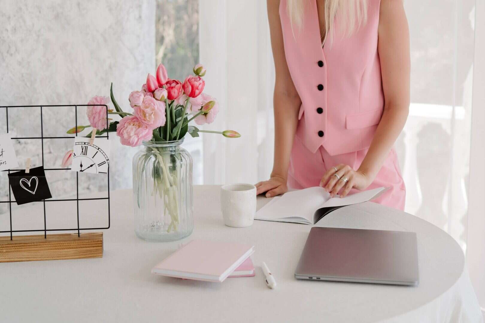 Workspace with notebooks, laptop, coffee mug, and flowers while a woman in pink reviews a planner. Represents the conclusion of a hybrid planner system, highlighting organization, balance, and productivity.
