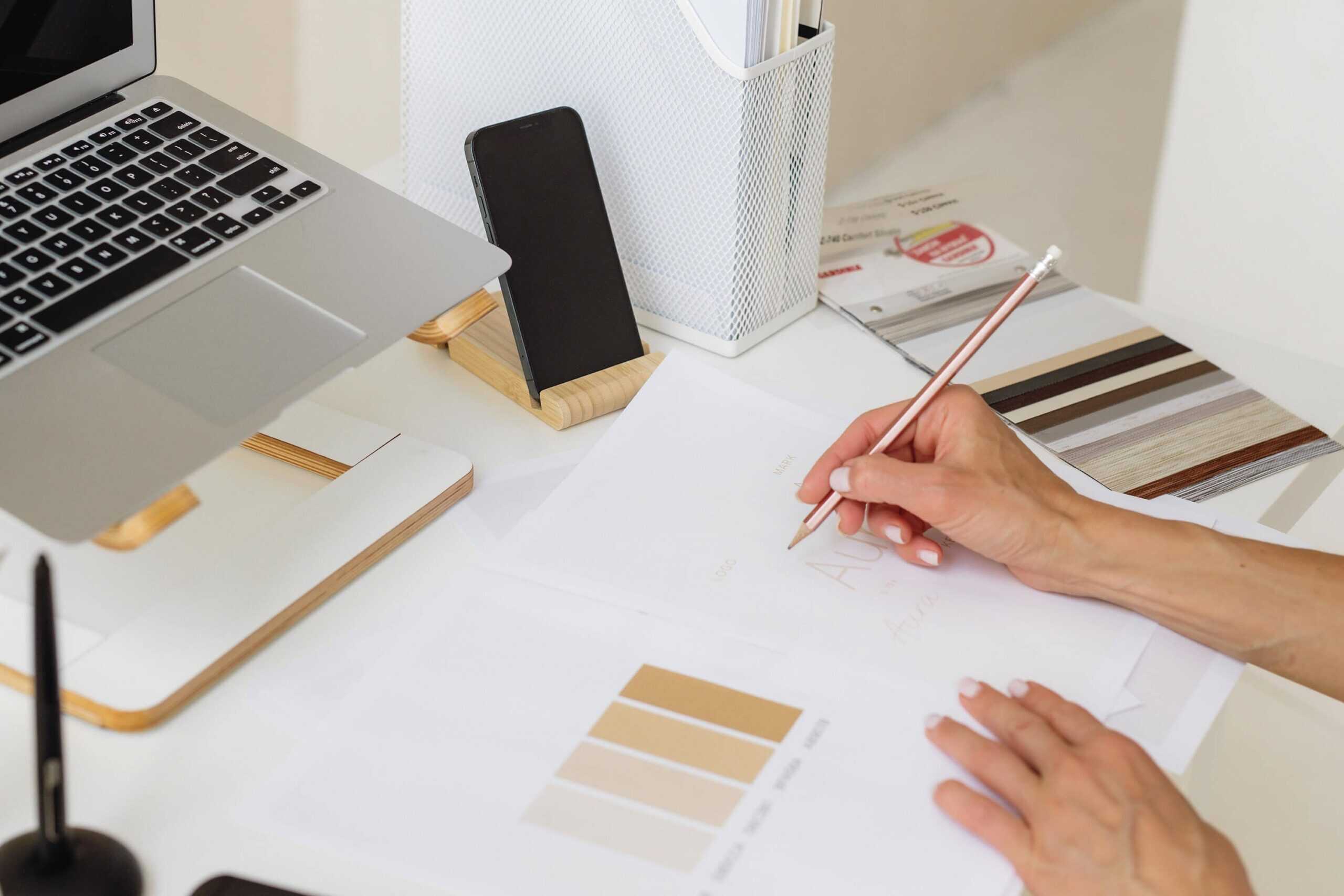 Hybrid planner system setup with a laptop on a stand, smartphone, and a person writing on printable planner pages with a pencil. Shows how digital and printable planners can be used together for an organized workflow.