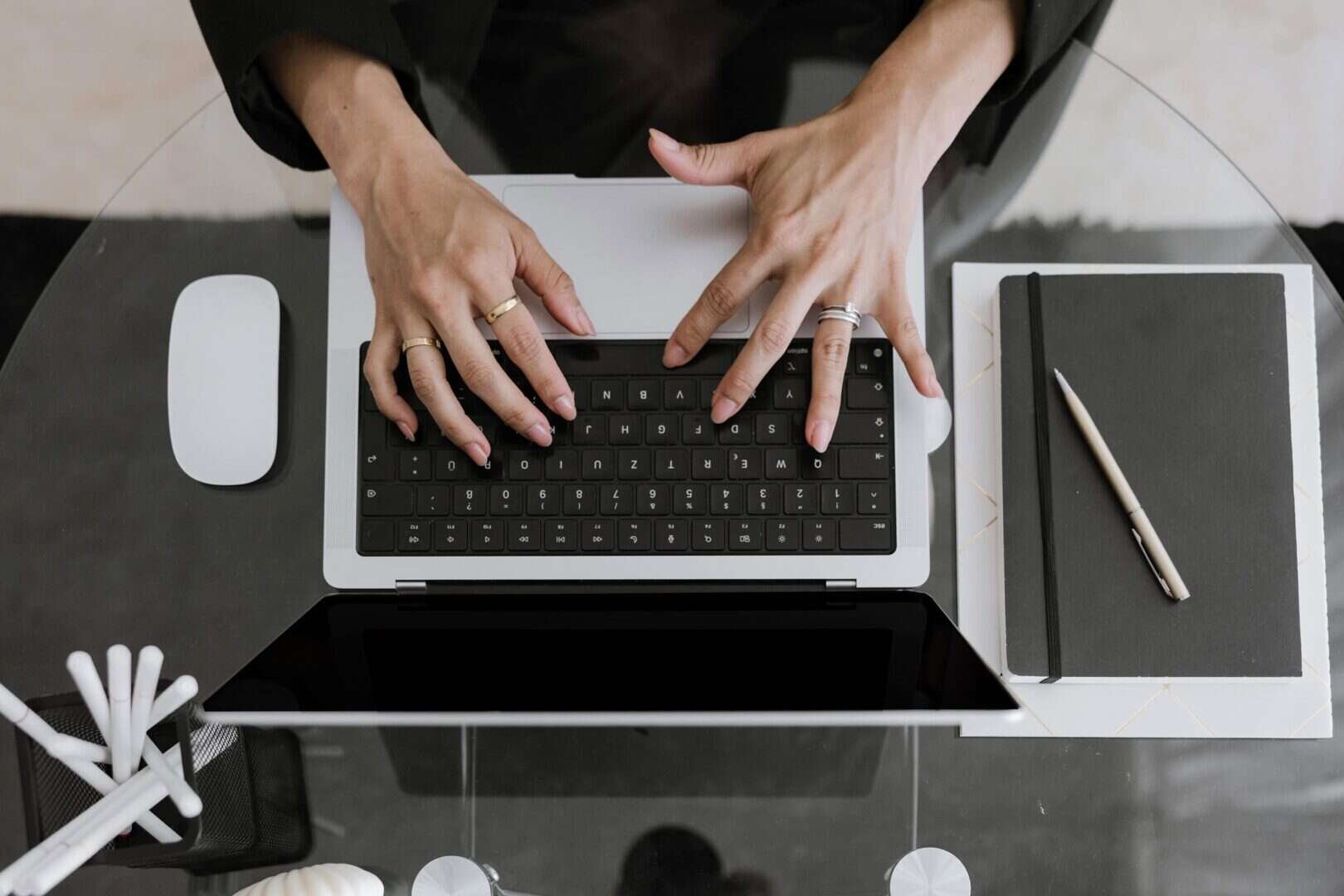 Workspace with a laptop, notebook, and pen on a glass desk while hands type on the keyboard. Represents hybrid daily planning for entrepreneurs using both printable planners for brainstorming and digital tools for tasks.