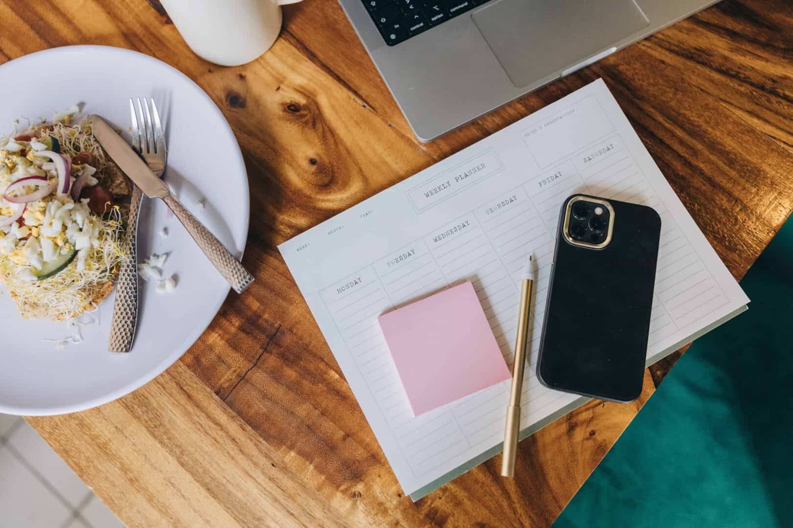 Weekly planner on a wooden table with a smartphone, gold pen, sticky notes, and a plate of food beside a laptop. Represents hybrid planning for moms who balance meal planning and family schedules with digital and printable planners.