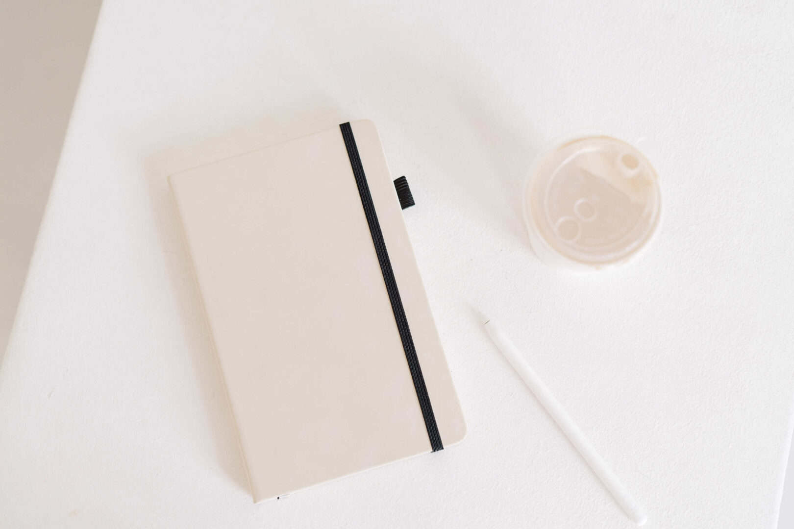 Minimalist flat lay of a closed beige planner notebook with a black elastic band, a white stylus pen, and a coffee cup on a white desk. Represents the simplicity and clarity that printable or digital planners can bring to creative entrepreneurs and stay-at-home moms balancing daily routines and personal projects.