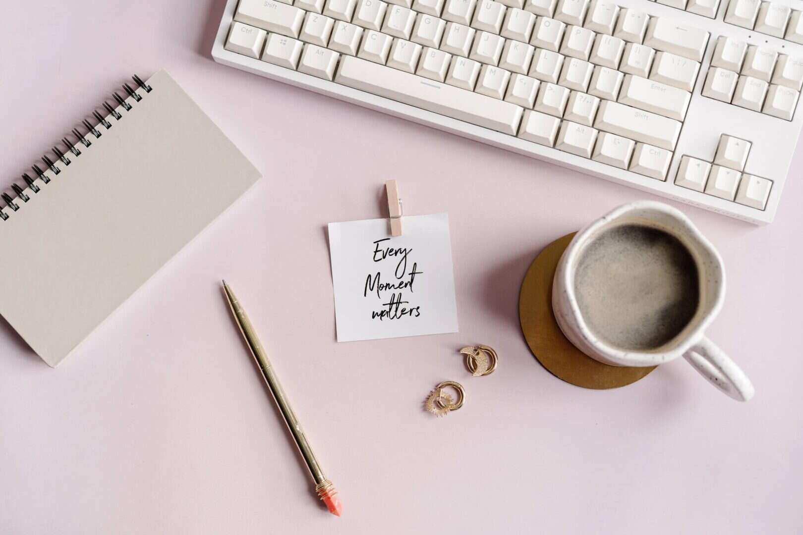Flat lay of a stylish planner desk setup with a spiral notebook, gold pen, motivational card reading “Every Moment Matters,” a cup of coffee, gold earrings, and a white keyboard on a soft pink surface. Ideal visual for introducing a guide to the best printable planners, digital planner systems, and planner tools for moms and entrepreneurs.