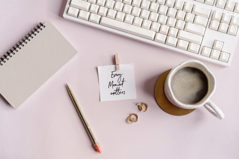 Flat lay of a stylish planner desk setup with a spiral notebook, gold pen, motivational card reading “Every Moment Matters,” a cup of coffee, gold earrings, and a white keyboard on a soft pink surface. Ideal visual for introducing a guide to the best printable planners, digital planner systems, and planner tools for moms and entrepreneurs.