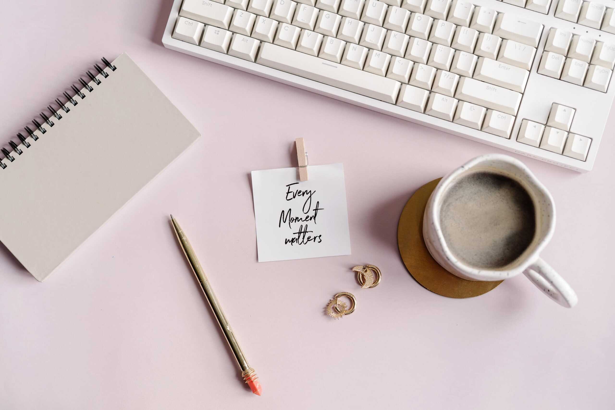 Flat lay of a stylish planner desk setup with a spiral notebook, gold pen, motivational card reading “Every Moment Matters,” a cup of coffee, gold earrings, and a white keyboard on a soft pink surface. Ideal visual for introducing a guide to the best printable planners, digital planner systems, and planner tools for moms and entrepreneurs.