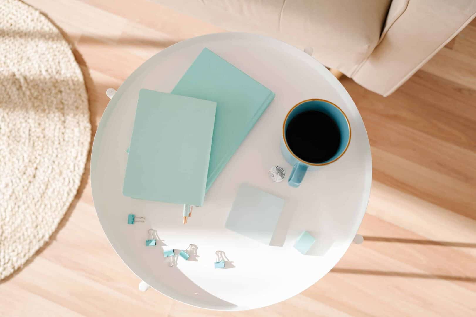 Top view of a white table with aqua notebooks, sticky notes, binder clips, and a coffee mug. Represents the tools and habits that make a planner routine stick for daily and weekly planning.