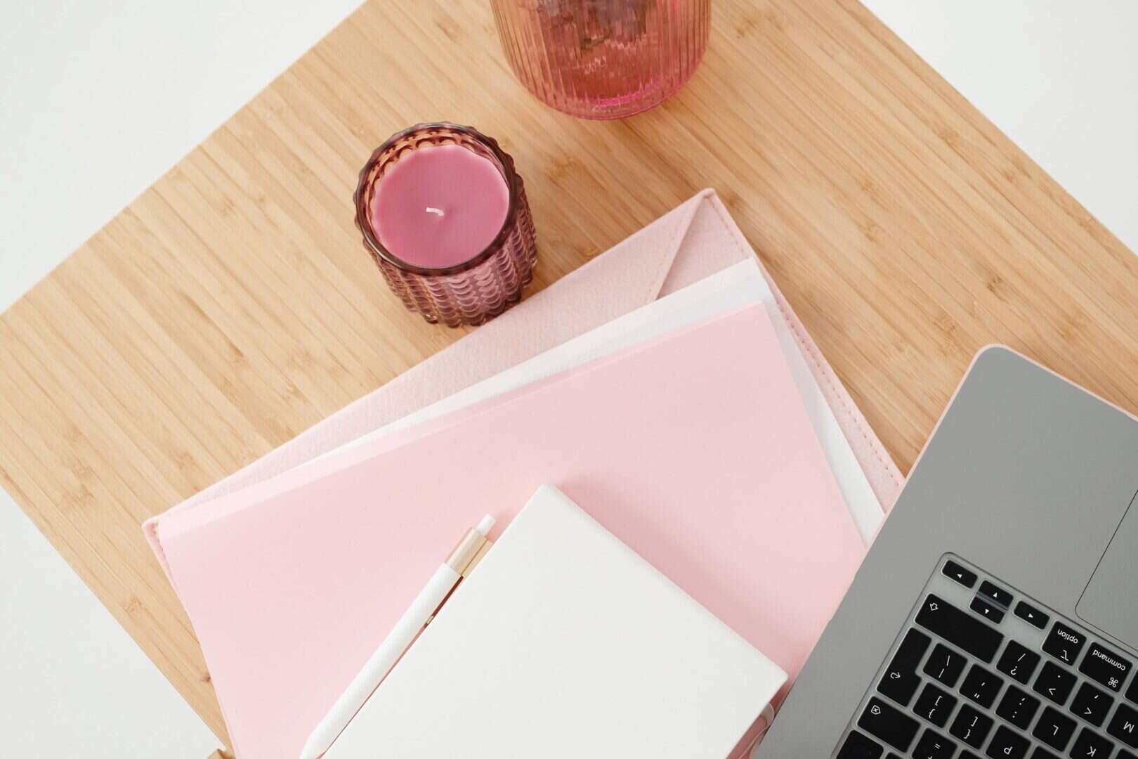 Pink planner with a pen, laptop, and candle on a wooden desk. Represents the tools and environment that help create a planner routine that sticks.