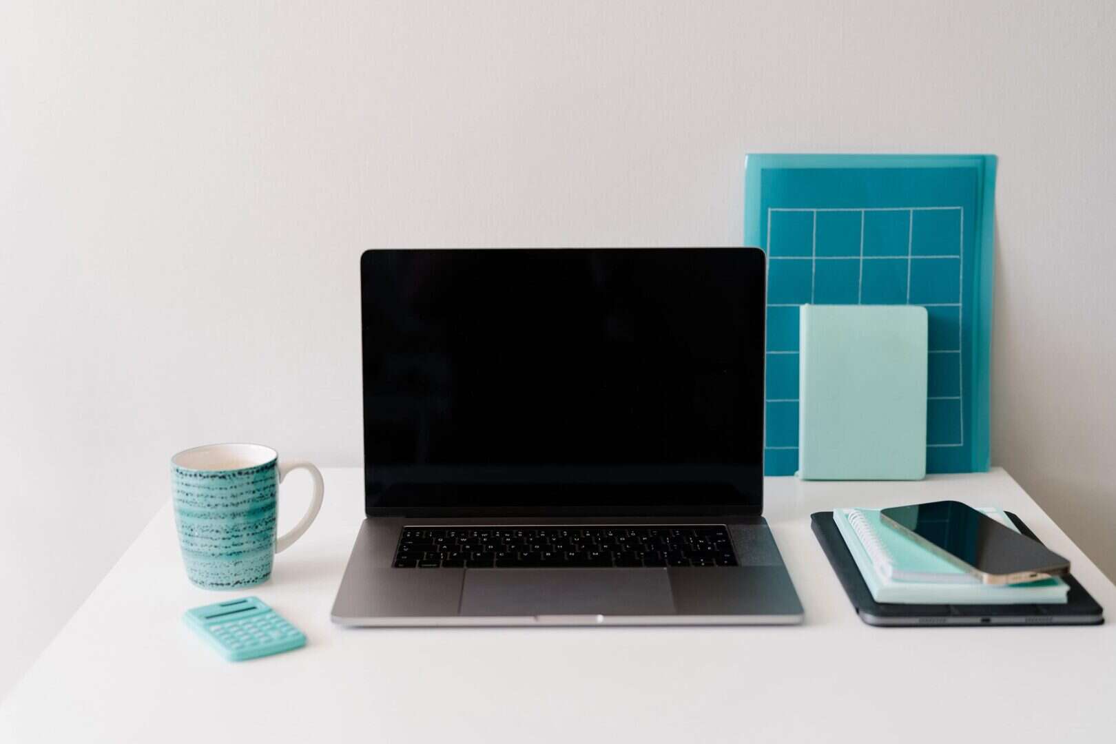 Organized workspace with a laptop, smartphone, spiral notebook, turquoise planner, wall calendar, calculator, and coffee mug on a white desk. Represents essential planner tools and resources that support productivity and organization for moms and entrepreneurs.