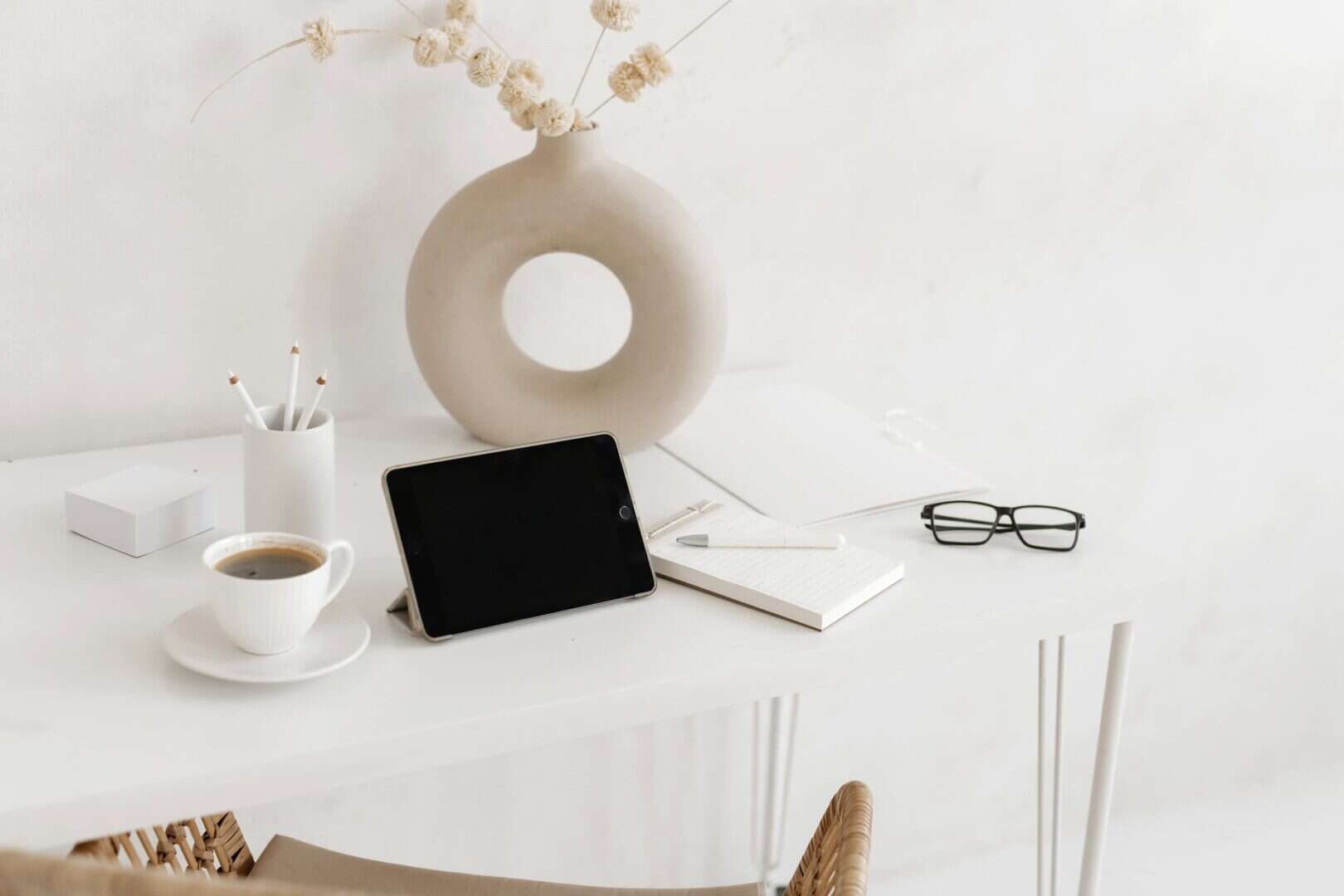 Minimalist home office setup with a tablet, lined notepad, white pen, coffee cup, eyeglasses, and neutral decor on a white desk. Reflects an organized workspace where printable planners and essential planning tools support focus and productivity for moms and entrepreneurs.